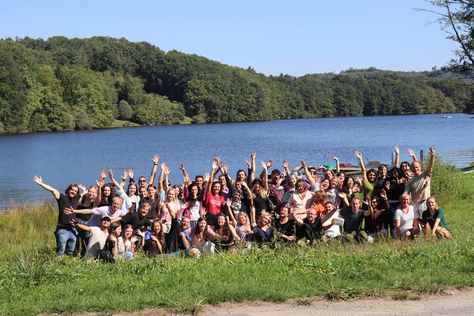 Around sixty participants of the Portraits of the Soul youth exchange, arms raised, in front of a lake in France